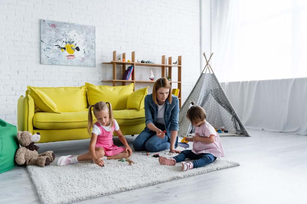 воспитательница детского сада сидит на ковре и играет с девочкой с синдромом Дауна рядом с ребенком в игровой комнате  - Фото, изображение