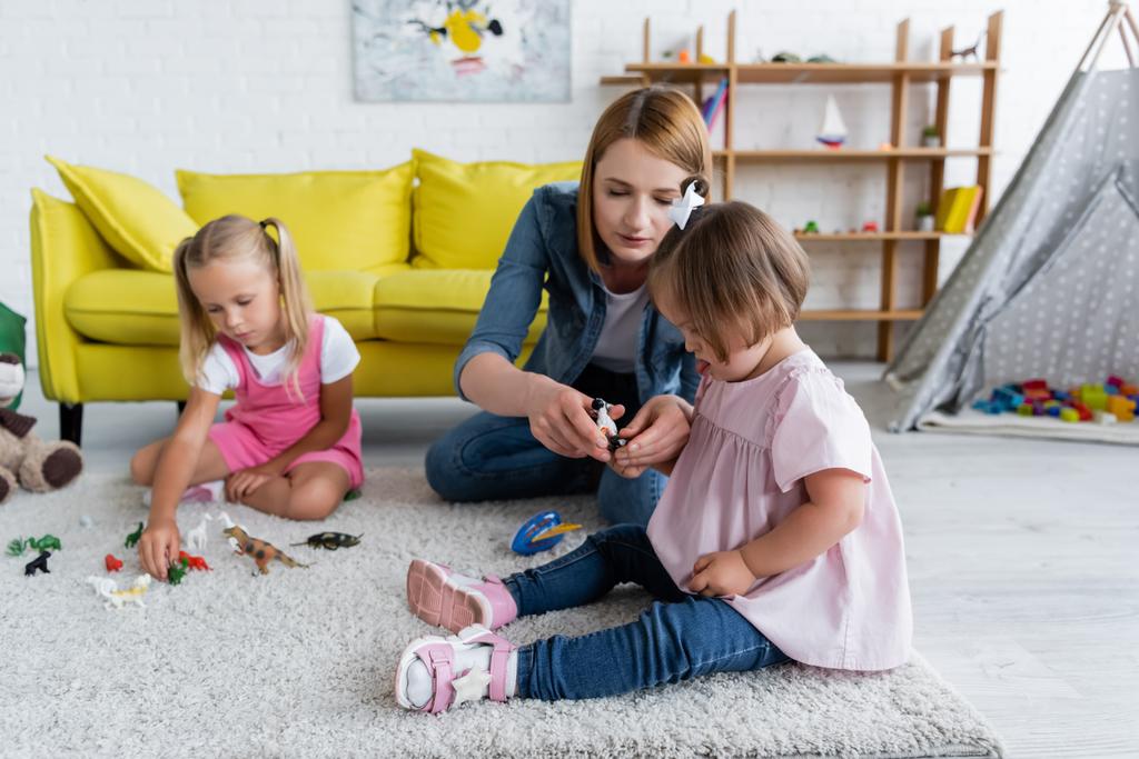 вчитель дитячого садка дає іграшку дівчинці-малюкові з синдромом вниз поблизу розмитого дошкільника дитини в ігровій кімнаті
  - Фото, зображення
