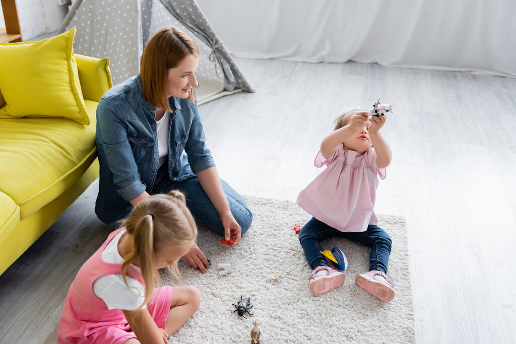 высокий угол зрения воспитателя детского сада глядя на малышку с синдромом Дауна, играющую с игрушкой рядом размытый ребенок дошкольного возраста в игровой комнате  - Фото, изображение