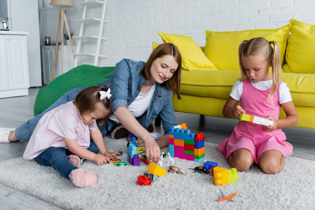 fröhliche Kindergärtnerin spielt Bausteine mit Vorschulmädchen und Kleinkind mit Down-Syndrom  - Foto, Bild