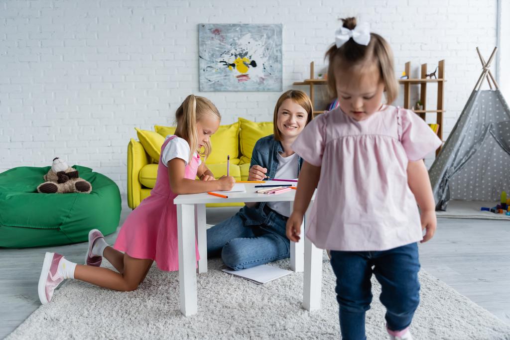 Улыбающийся воспитатель детского сада смотрит на размытого ребенка с синдромом Дауна, идущего в игровую комнату  - Фото, изображение