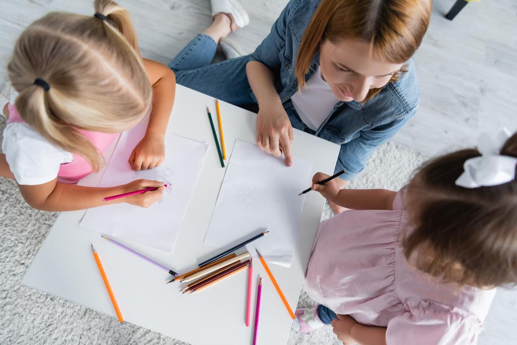 Kindergärtnerin sieht Kind mit Down-Syndrom in der Nähe von Vorschulmädchen auf Papier zeichnen - Foto, Bild