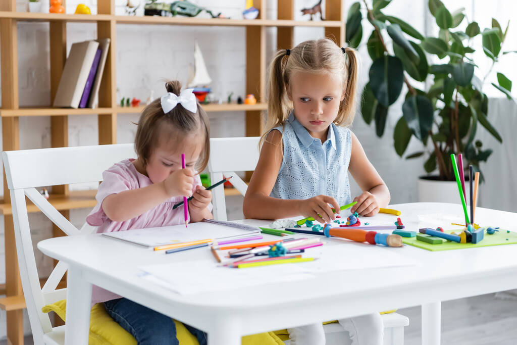 blonde girl looking at disabled toddler child with down syndrome drawing in private kindergarten  - Photo, Image