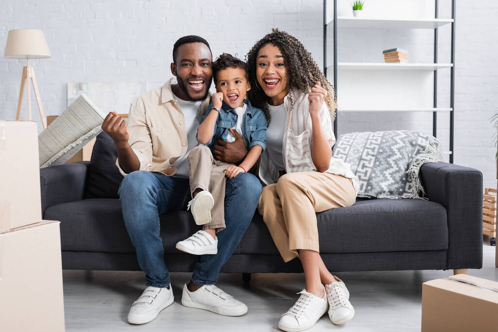 joyeuse famille afro-américaine montrant geste de triomphe sur le canapé dans un nouvel appartement - Photo, image