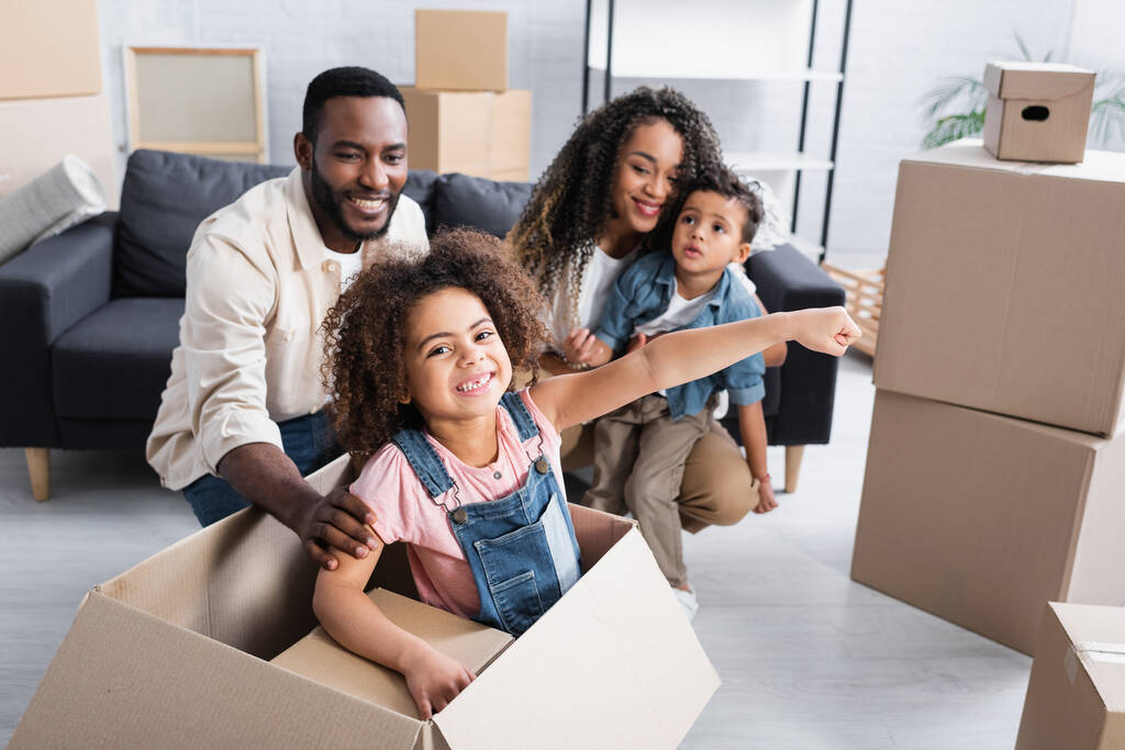 excitada chica afroamericana divirtiéndose en caja de cartón cerca de la familia en nuevo apartamento - Foto, Imagen