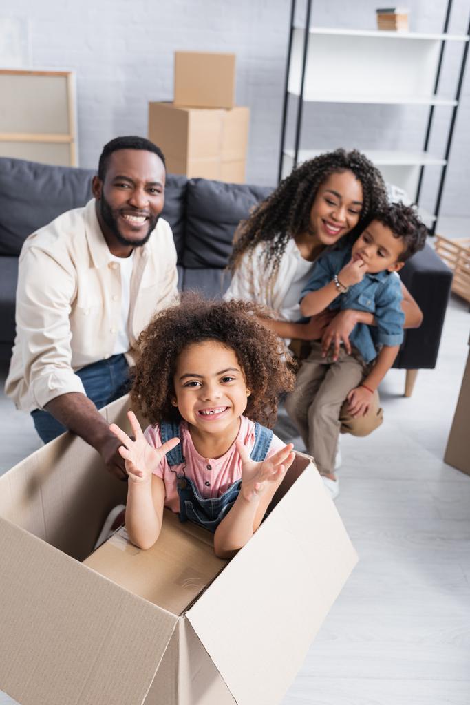 Afrykańska amerykańska dziewczyna w kartonowym pudełku pokazująca przerażający gest w pobliżu rozmytej rodziny w nowym mieszkaniu - Zdjęcie, obraz
