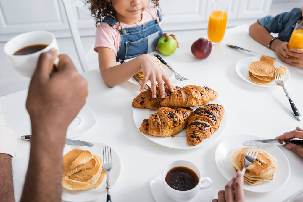 vista recortada de chica afroamericana tomando croissant durante el desayuno con la familia borrosa - Foto, imagen