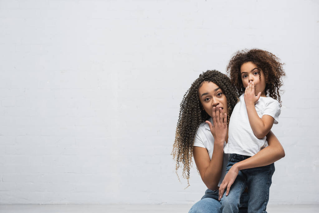 schockierte afrikanisch-amerikanische Frau mit Mädchen, das Mund mit der Hand bedeckt, während sie in die Kamera auf grau schaut - Foto, Bild