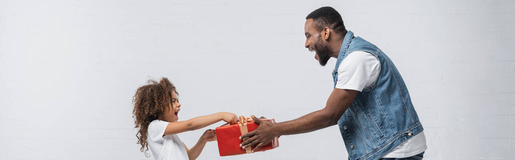 Afro-amerikan bir adamın, gri pankartta, afişte, şaşırmış kızına hediyesini sunuşunun yan görüntüsü. - Fotoğraf, Görsel