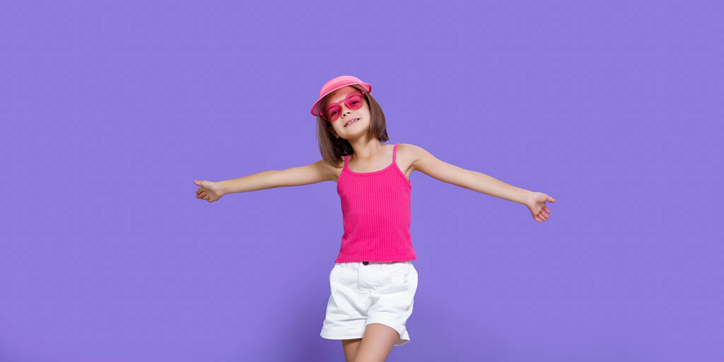 Κοριτσάκι με λευκό σορτς, ροζ μπλουζάκι, ροζ μοντέρνα γυαλιά και καλοκαιρινό γείσο που ποζάρει με τα χέρια στο πλάι σε μωβ φόντο στο στούντιο. - Φωτογραφία, εικόνα