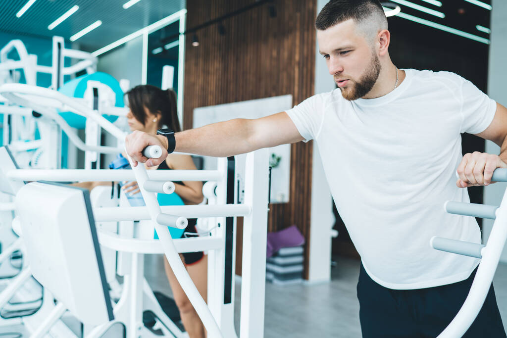 Geconcentreerde man met baard en trendy kapsel staand op een elliptische machine en cardio-oefeningen tijdens de training in een moderne lichte fitnessruimte - Foto, afbeelding