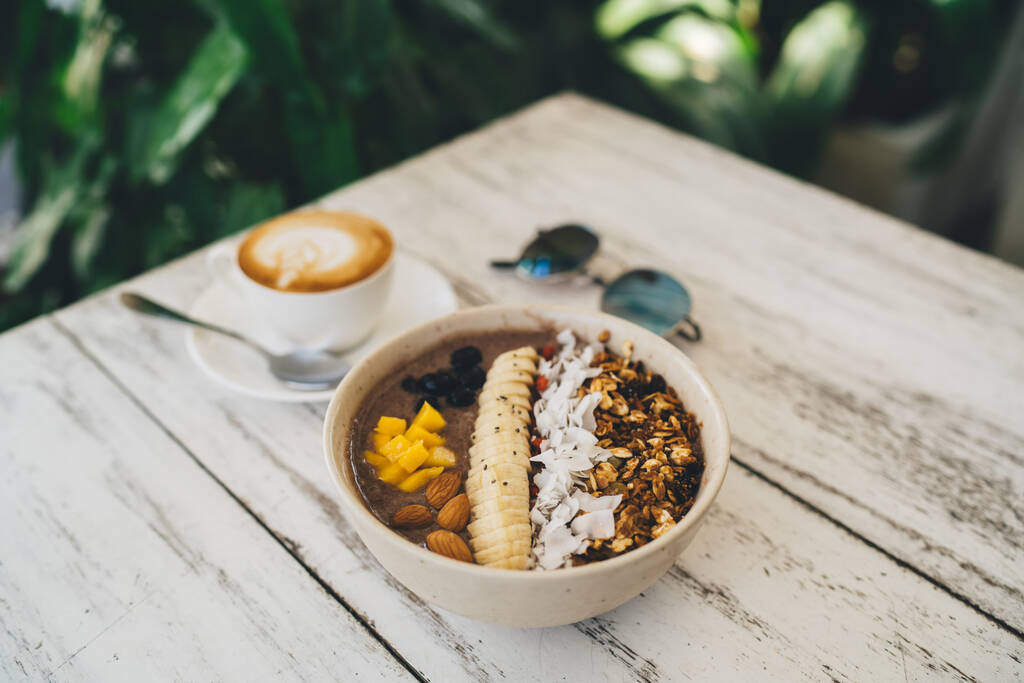Wysoki kąt apetyczny granola i plasterki banana z orzechami w pobliżu filiżanki aromatycznego cappuccino na drewnianym stole w kawiarni - Zdjęcie, obraz