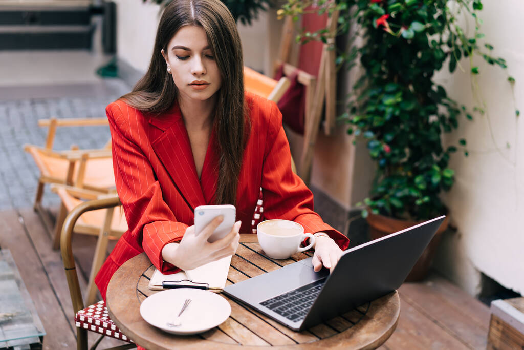 Mujer joven en traje rojo elegante usando teléfono inteligente y portátil para el trabajo de forma remota mientras está sentado en la terraza de la cafetería con taza de café - Foto, Imagen