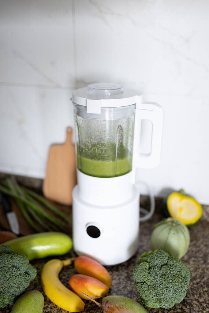 Świeże składniki dla zdrowego detox koktajl zielony kolor. Mikser z smoothie na dietę na stole kuchennym, obok leżących brokułów i innych warzyw i owoców. - Zdjęcie, obraz