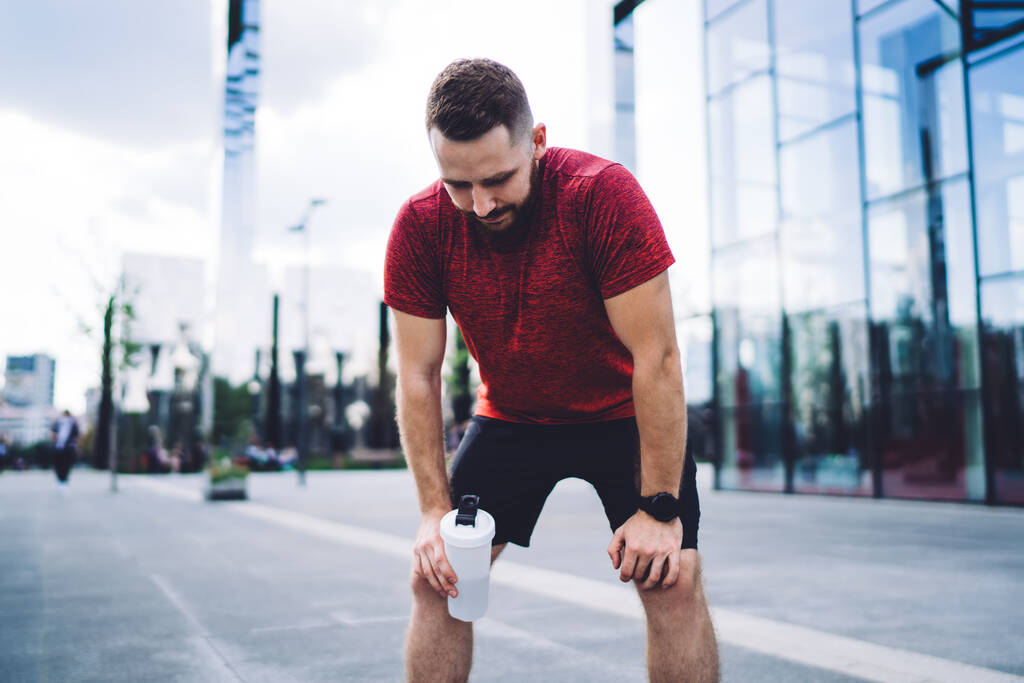 Stanco jogger maschile in maglietta rossa con bottiglia in mano in piedi vicino al centro durante la pausa durante il riposo dopo l'allenamento - Foto, immagini