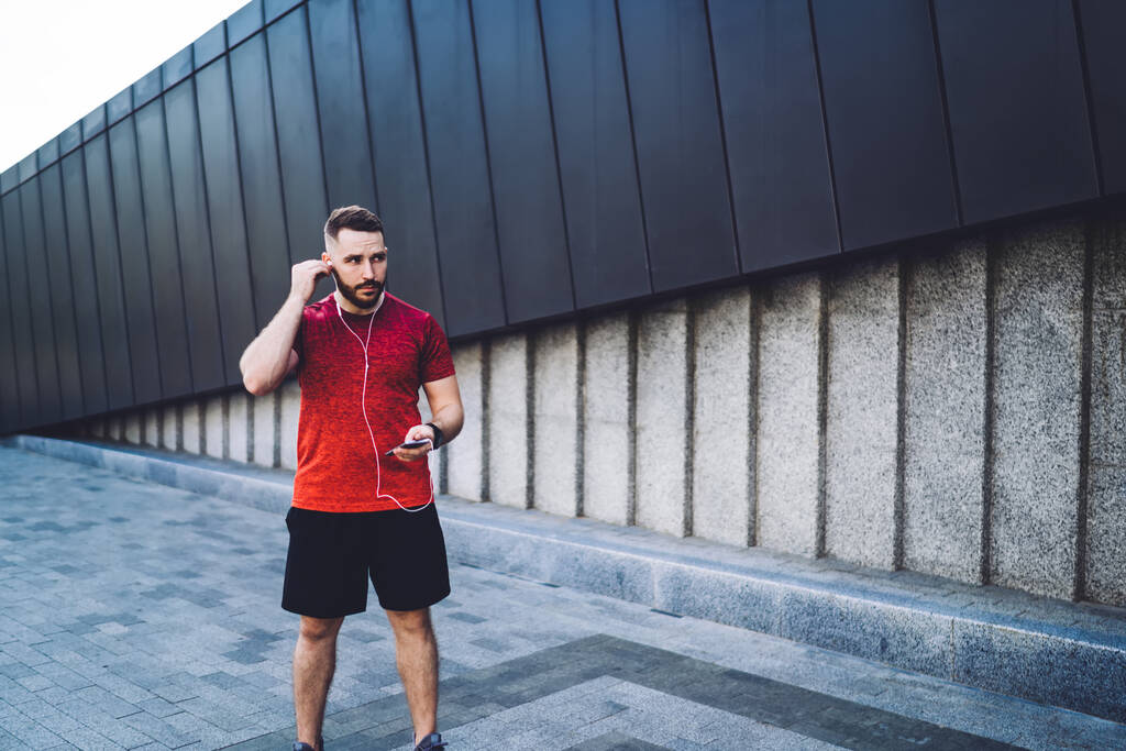 Soustředěný vousatý sportovec ve sportovním oblečení ve sluchátkách a poslouchající písně při stání na chodníku u moderní budovy ve městě - Fotografie, Obrázek