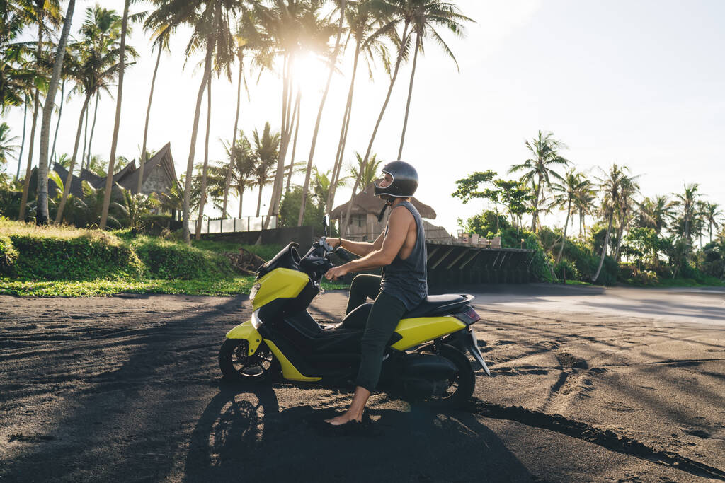 Vue latérale du motard masculin pieds nus méconnaissable en vêtements décontractés et casque à moto moderne sur le rivage sablonneux pendant les vacances d'été - Photo, image