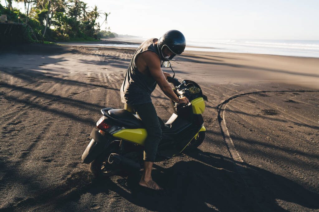 Boční pohled plné tělo sportovní muž biker v helmě stojící s moderní motocykl na písečné pobřeží v slunné exotické zemi - Fotografie, Obrázek