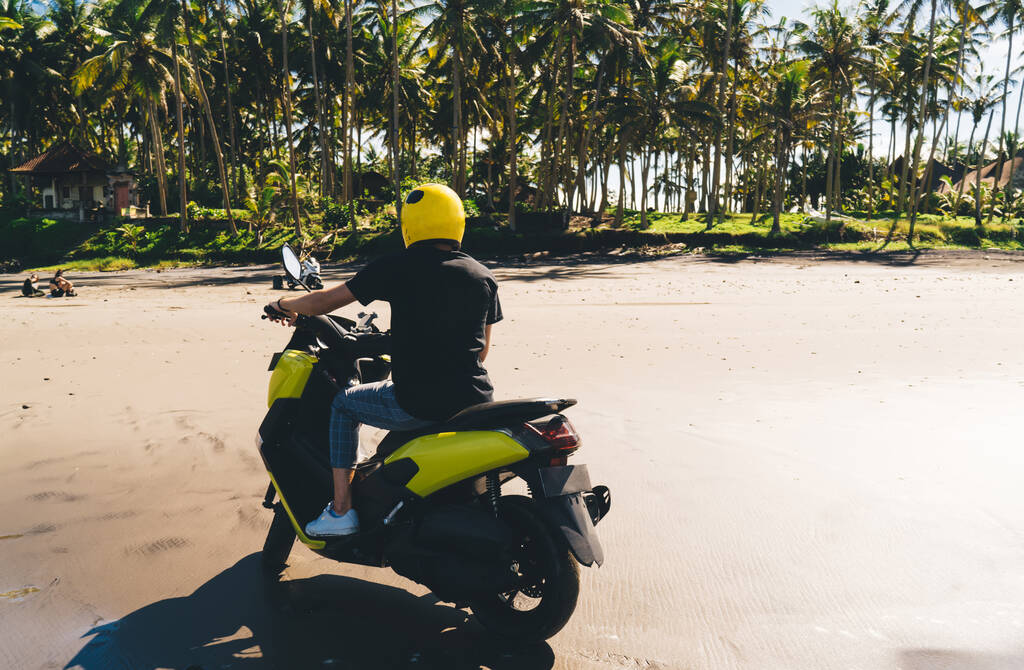 Achteraanzicht van anonieme persoon in casual kleding en helm rijden motor op zandstrand tegen tropisch landschap op zonnige dag - Foto, afbeelding