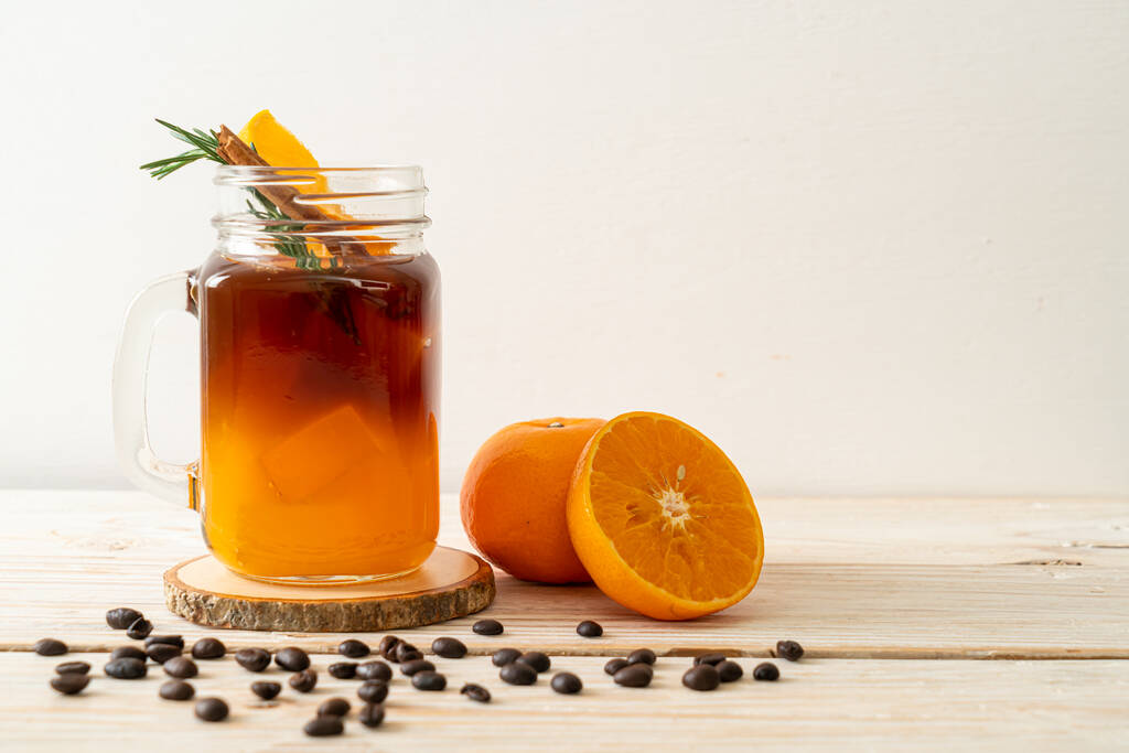 Sklenice ledové americké černé kávy a vrstva pomerančové a citrónové šťávy zdobené rozmarýnem a skořicí na dřevěném pozadí - Fotografie, Obrázek