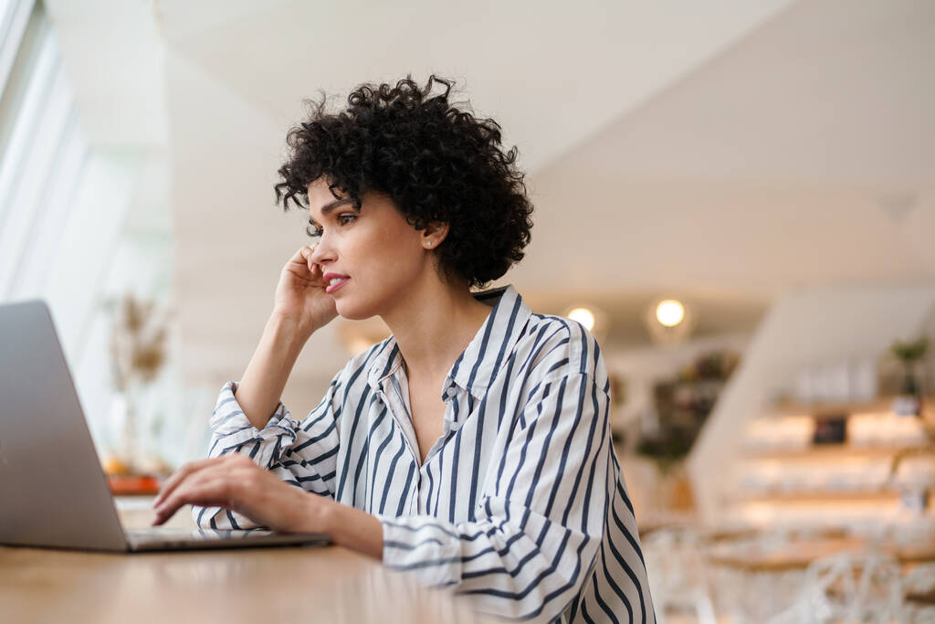 Kaunis keskittynyt kihara nainen työskentelee kannettavan tietokoneen kanssa istuessaan kahvilassa - Valokuva, kuva