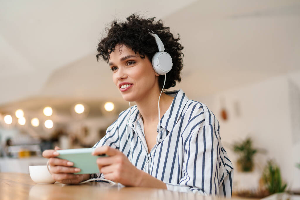 Mulher alegre agradável em fones de ouvido usando telefone celular enquanto bebe café no café - Foto, Imagem