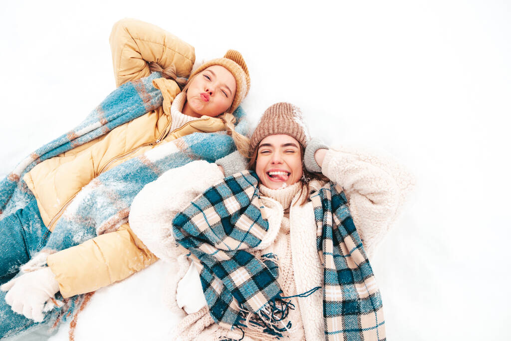 Dwie młode, piękne, uśmiechnięte hipsterki w modnych ciepłych ubraniach i szalikach. Beztroskie kobiety pozujące na ulicy w parku. Pozytywne czyste modele leżące w śniegu. Cieszymy się zimowymi chwilami. Koncepcja świąteczna - Zdjęcie, obraz
