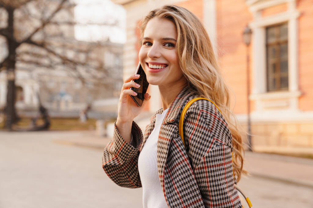 Sonriente joven rubia hablando por teléfono móvil mientras está de pie en una calle de la ciudad - Foto, imagen