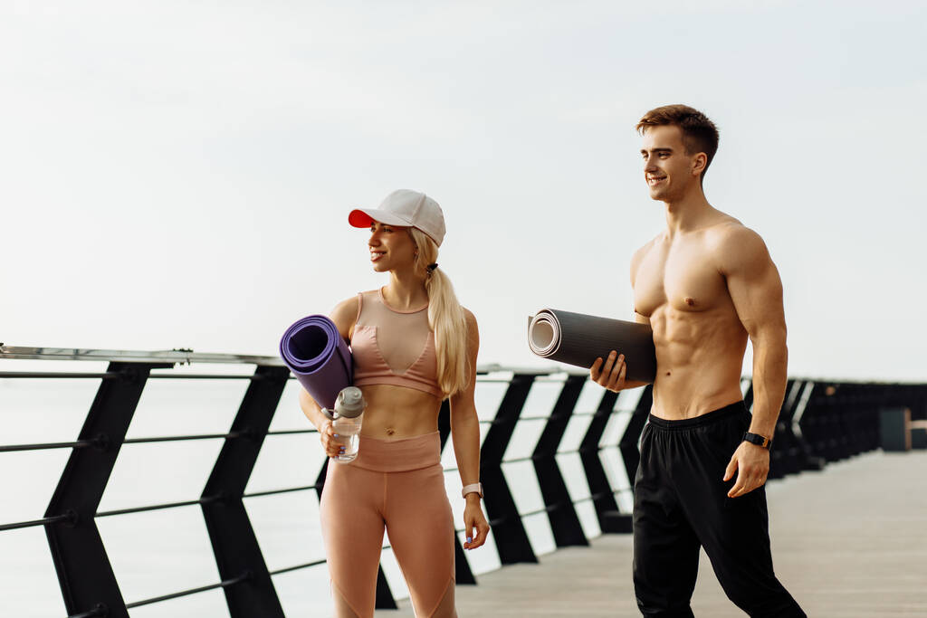 Jonge fitnesspaar gaan naar de training, man en vrouw met fitnessmatten in handen, klaar om buiten te trainen op de pier, sport en gezonde levensstijl - Foto, afbeelding