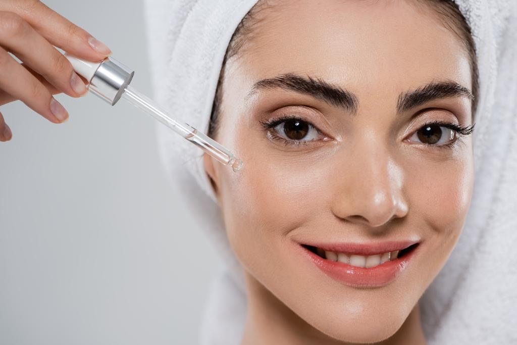 close up of happy young woman applying moisturizing serum with pipette isolated on grey - Photo, Image