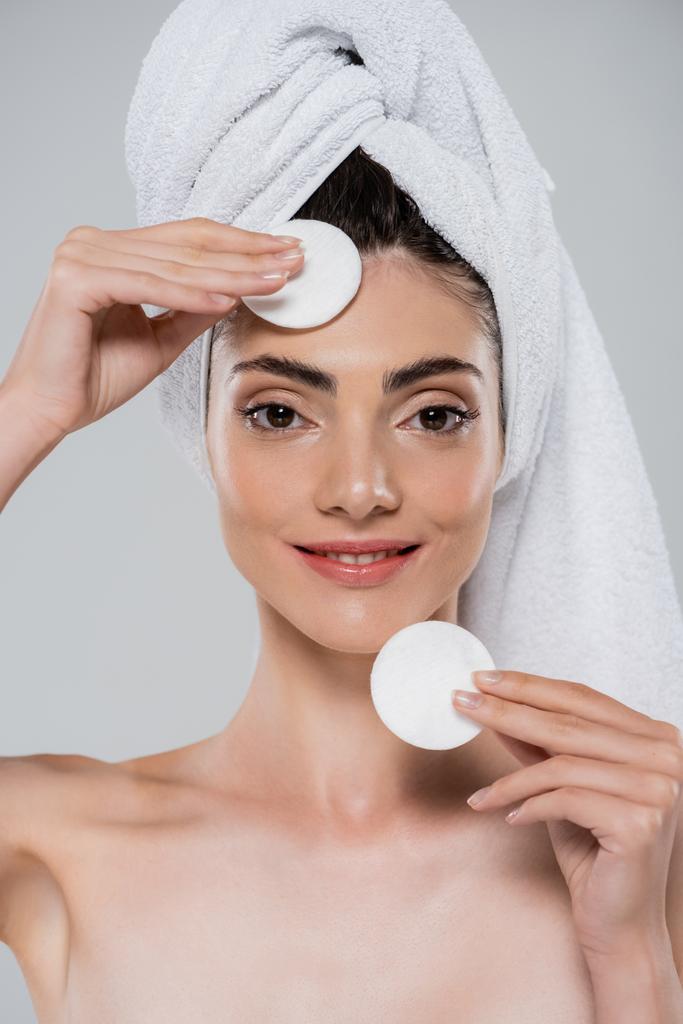 smiling woman with towel on head removing makeup with cotton pads isolated on grey - Photo, Image