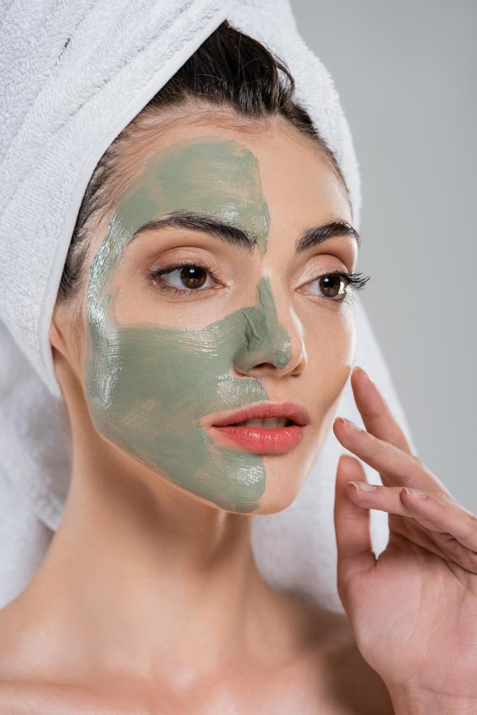 junge Frau mit Handtuch auf dem Kopf und grüner Lehmmaske im Gesicht isoliert auf grau - Foto, Bild