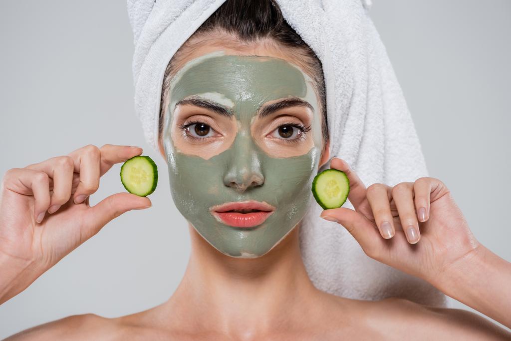 mladá žena se zelenou hliněnou maskou na obličeji držící nakrájenou okurku izolovanou na šedé - Fotografie, Obrázek