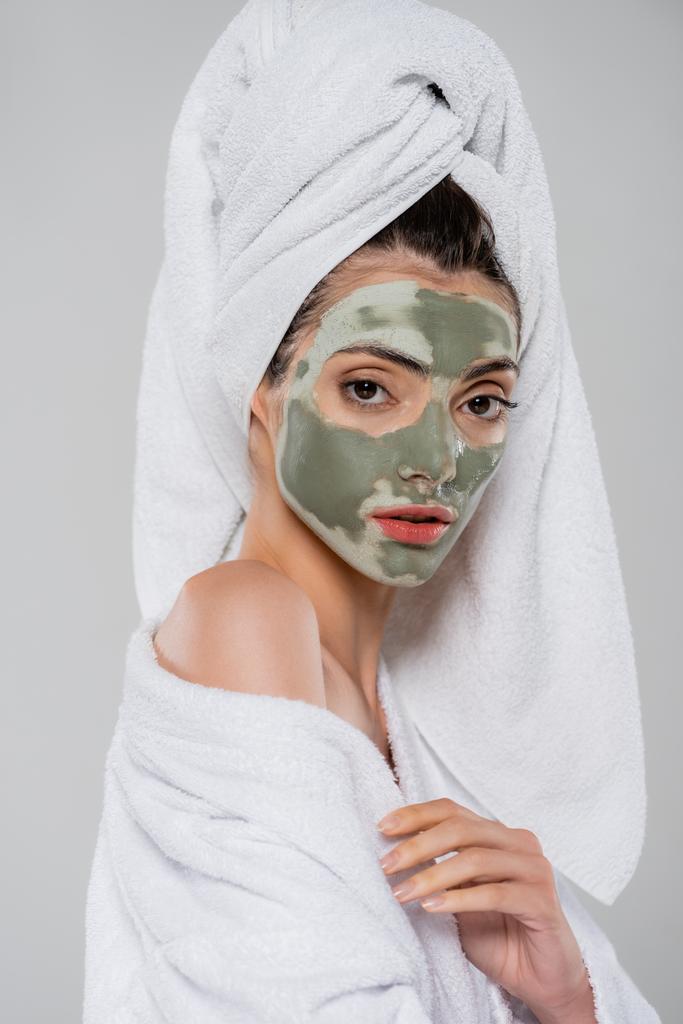 młoda kobieta z ręcznikiem na głowie i glinianą maską na twarzy patrząc na kamerę odizolowaną na szaro - Zdjęcie, obraz
