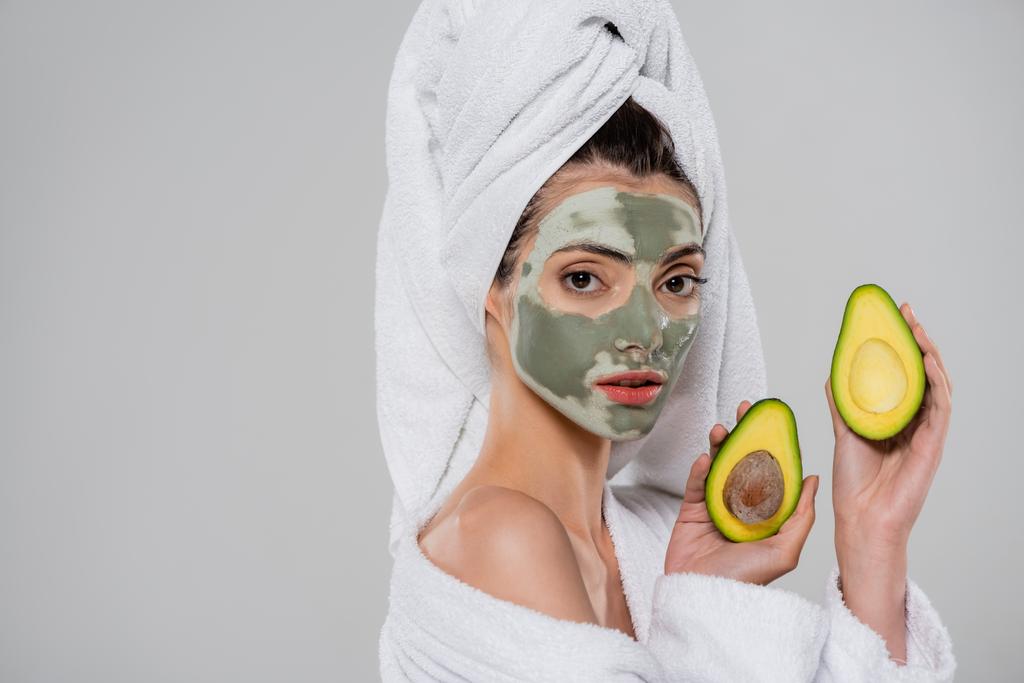 молодая женщина с полотенцем на голове и зеленой глиняной маской держа половинки авокадо изолированы на сером - Фото, изображение