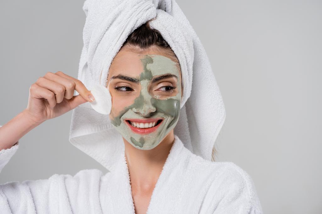 gelukkig jong vrouw in badjas verwijderen klei masker met katoen pad geïsoleerd op grijs  - Foto, afbeelding