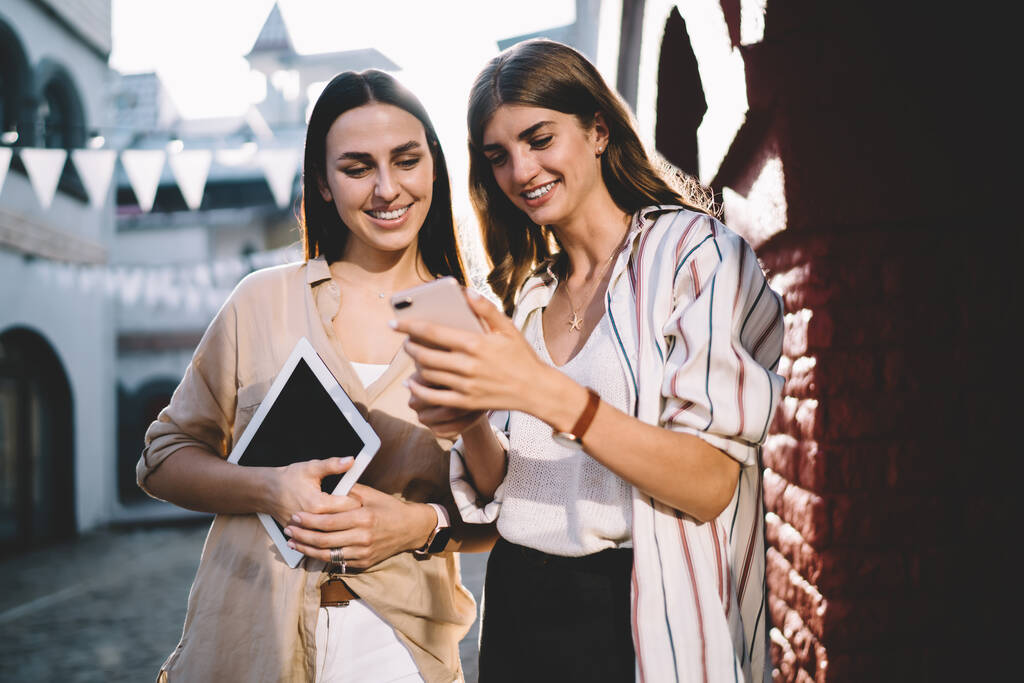 Glückliche weibliche Kollegen diskutieren lustige Inhalte während mobiler sozialer Netzwerke im urbanen Umfeld, fröhliche junge Frauen sehen positive Video-Vlog und lächeln bei freundlichen Treffen in der Stadt - Foto, Bild
