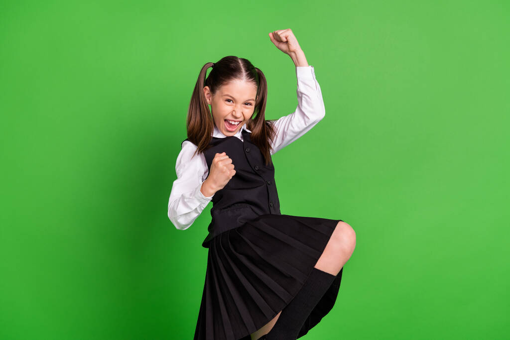 Photo of funny lucky school girl wear black white uniform rising fists smiling isolated green color background - Photo, Image