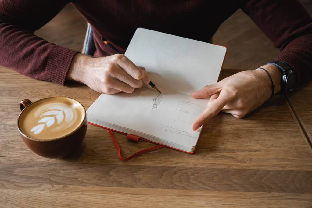 Ένας νεαρός άνδρας κάθεται στο καφέ, πίνει καφέ και κάνει σημειώσεις και σκίτσα στο σημειωματάριο. - Φωτογραφία, εικόνα