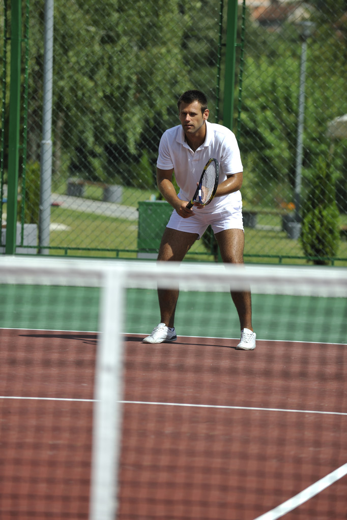 Молодой человек играет в теннис на открытом воздухе на оранжевом теннисном поле рано утром
 - Фото, изображение
