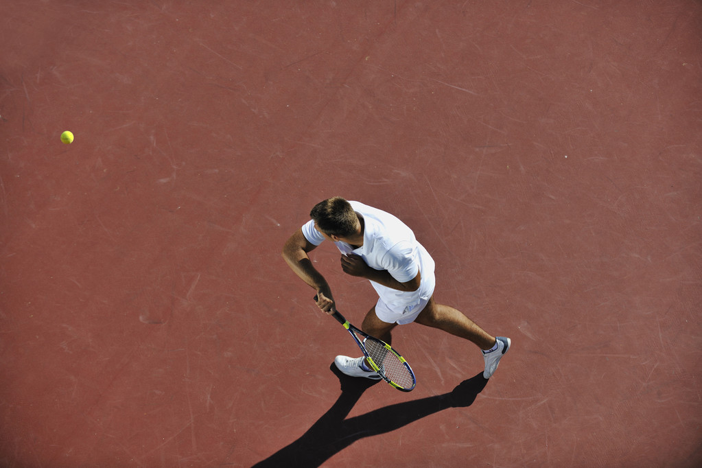 Junger Mann spielt frühmorgens Tennis im Freien auf orangefarbenem Tennisplatz - Foto, Bild