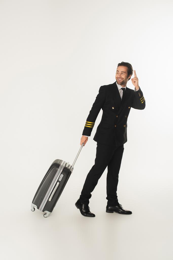 Glimlachende piloot met koffer wijzend met vinger op witte achtergrond  - Foto, afbeelding