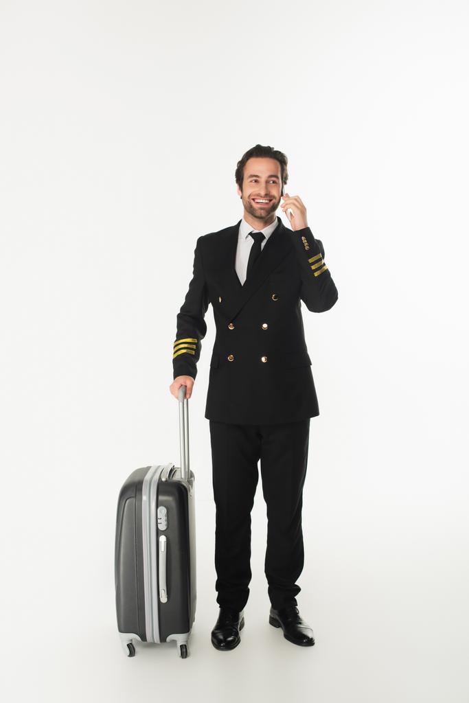 Mosolygós pilóta beszél a mobiltelefon közelében bőrönd fehér háttér  - Fotó, kép