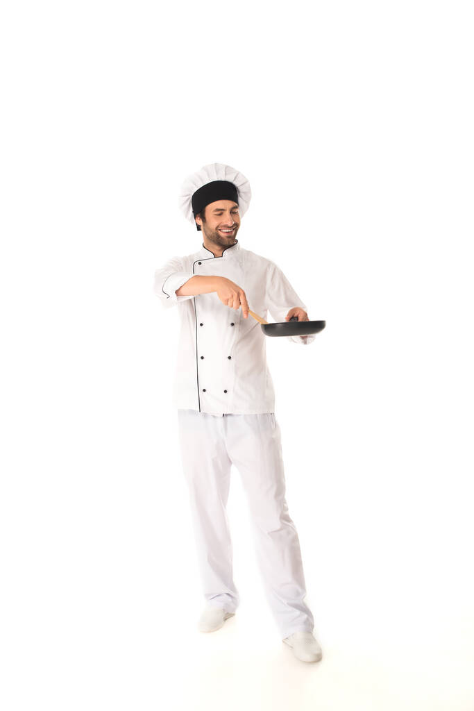 Volledige lengte van de lachende chef met koekenpan en houten spatel op witte achtergrond  - Foto, afbeelding