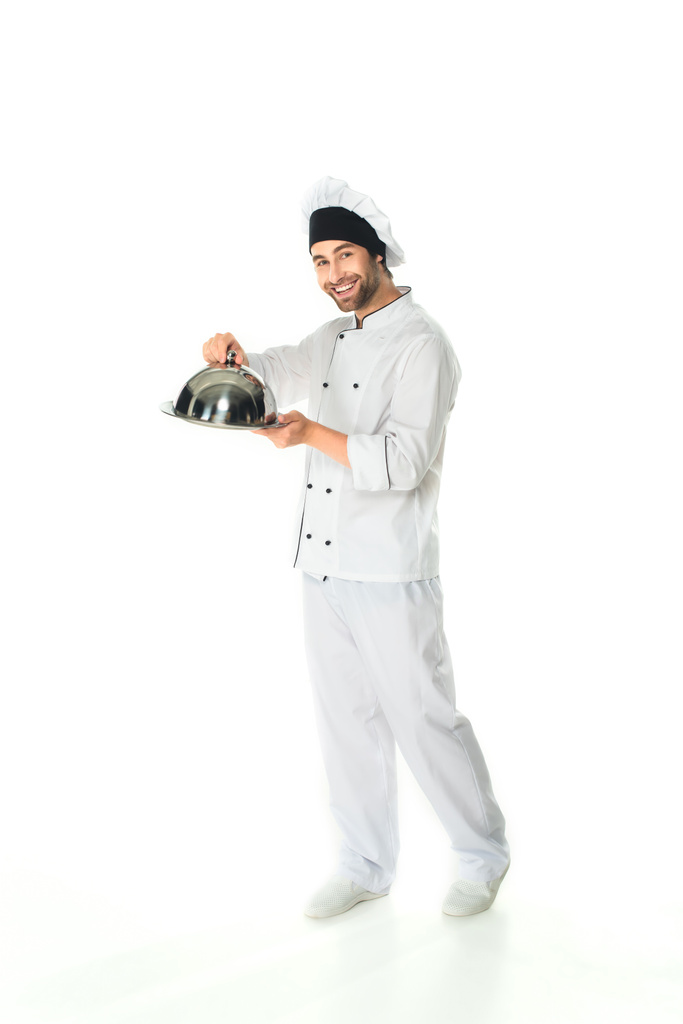 Longitud completa del chef sonriente sosteniendo la cloche y la bandeja sobre fondo blanco  - Foto, imagen