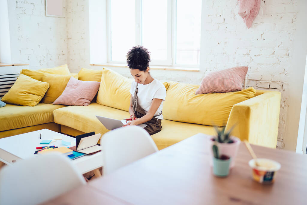 Donna afroamericana in abbigliamento casual con i capelli ricci che lavorano sul computer portatile mentre seduto sul divano giallo in soggiorno - Foto, immagini