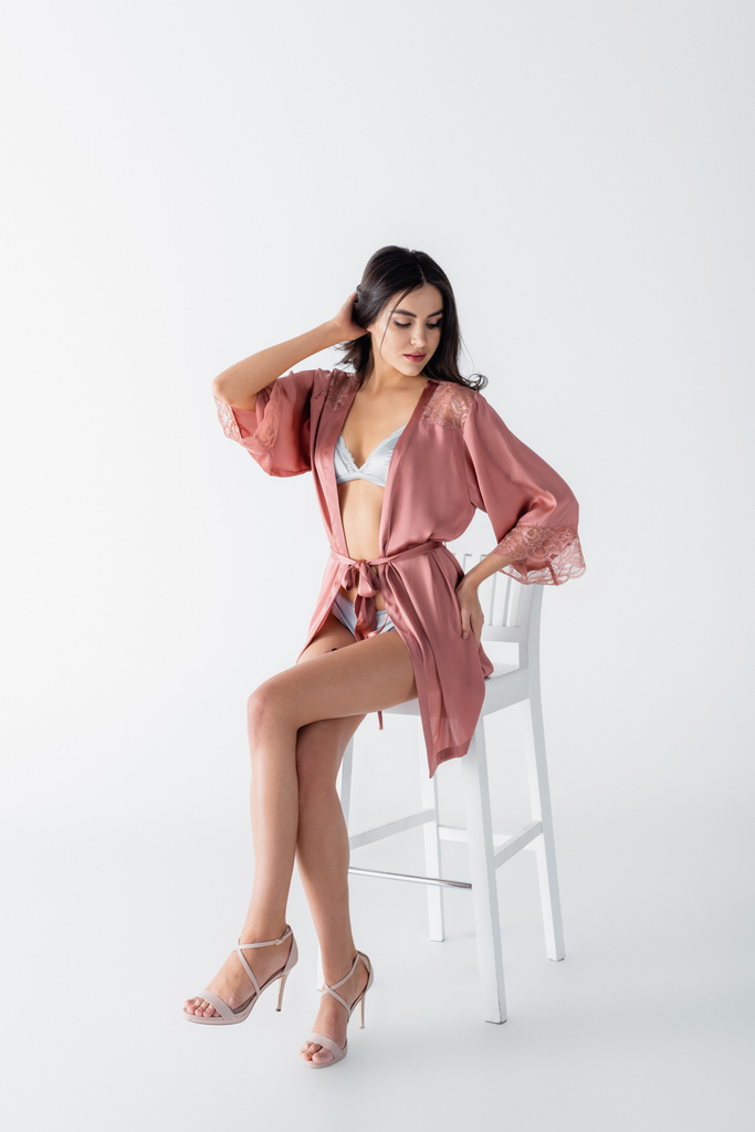 jonge vrouw in sexy ondergoed en satijnen mantel zitten op stoel op wit - Foto, afbeelding