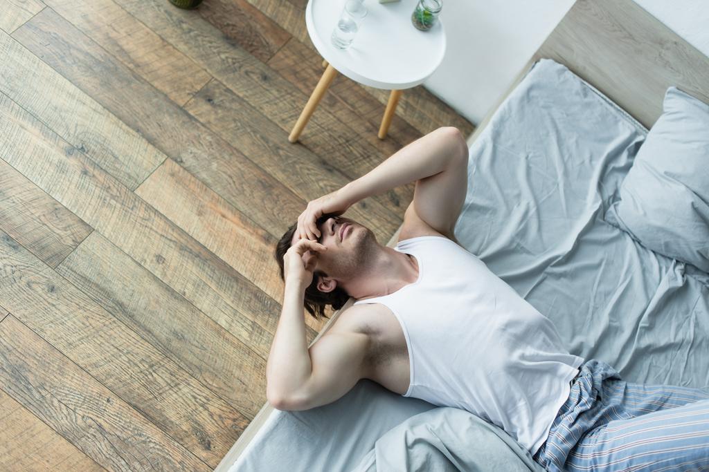 Hochwinkelaufnahme eines Mannes, der Augen mit Händen bedeckt, während er im Bett an Kater leidet - Foto, Bild