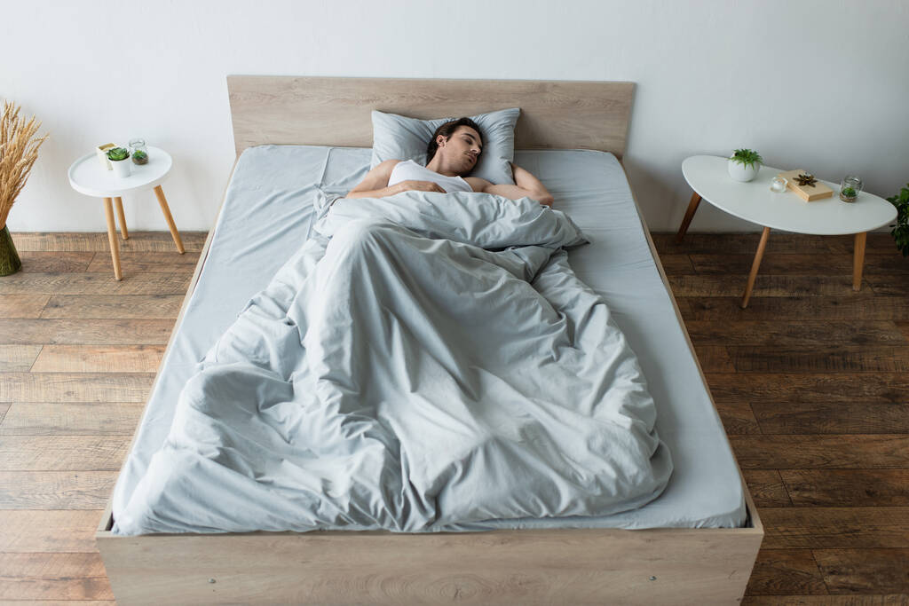 jonge man slapen op blauw beddengoed in de buurt van nachtkastjes - Foto, afbeelding