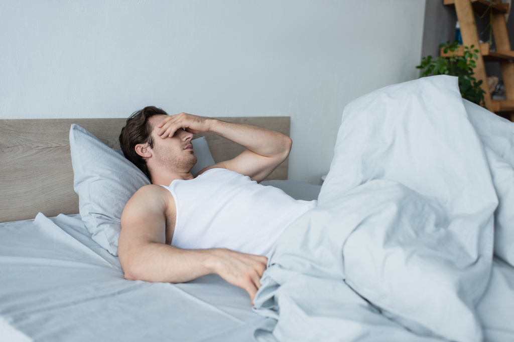 mladý muž zakrývající oči rukou při ležení v posteli - Fotografie, Obrázek